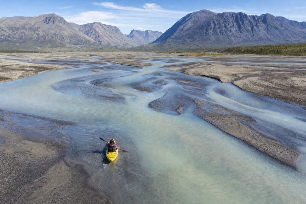 A kayaker reading the channels of a wide braided river below mountains
